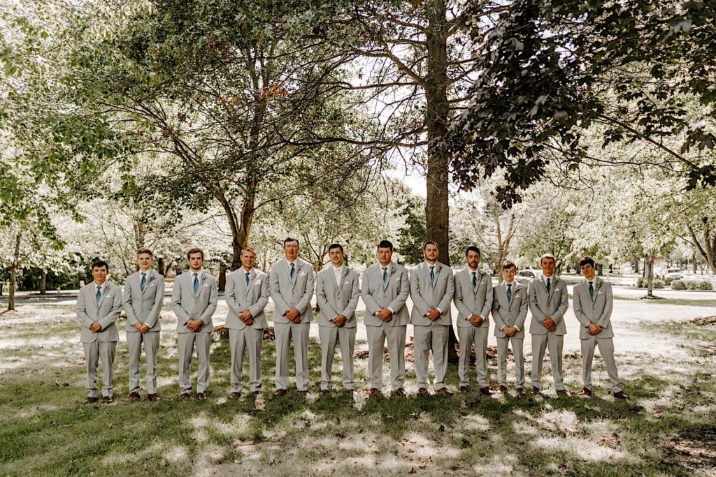 Groom standing in line with groomsmen all wearing grey suits in front of wooded area 