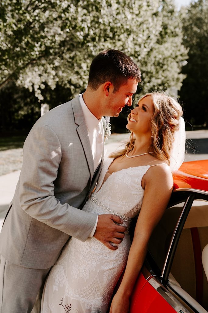 Bride leaning on red car with groom embracing her and looking into her eyes