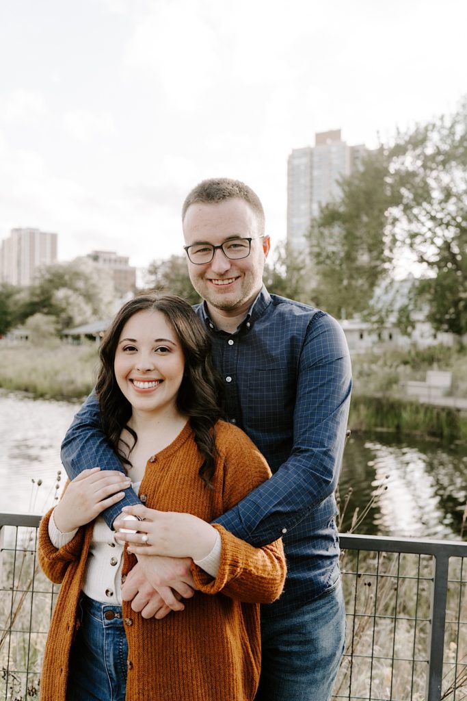 Man hugs his fiancée from behind while they both smile at the camera during their engagement session in Chicago's Lincoln Park