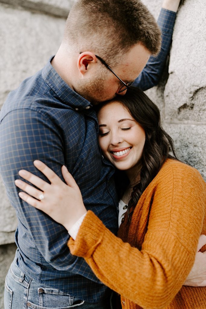 Man kisses his fiancée's head while she smiles in front of a brick wall
