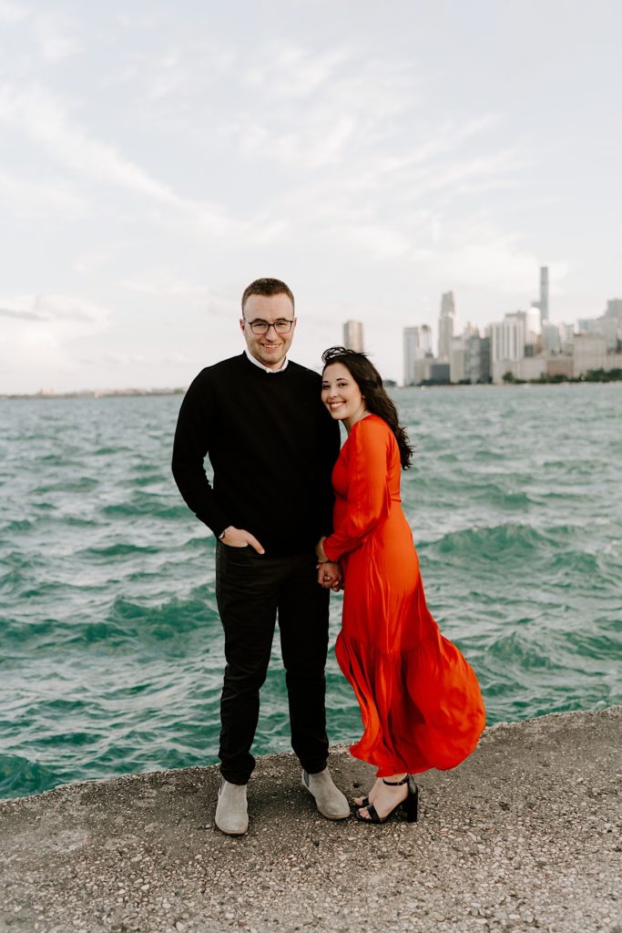 Couple pose next to one another and smile at the camera in front of the Chicago skyline and Lake Michigan at North Avenue Beach