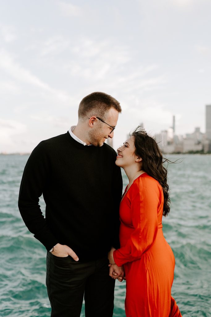 Couple pose next to one another and smile at one another in front of the Chicago skyline and Lake Michigan at North Avenue Beach