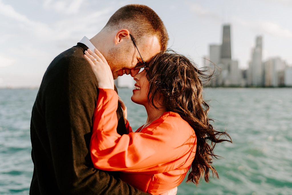 Couple smiling at one another while embracing and touching their foreheads together in front of the Chicago skyline and Lake Michigan during their engagement session