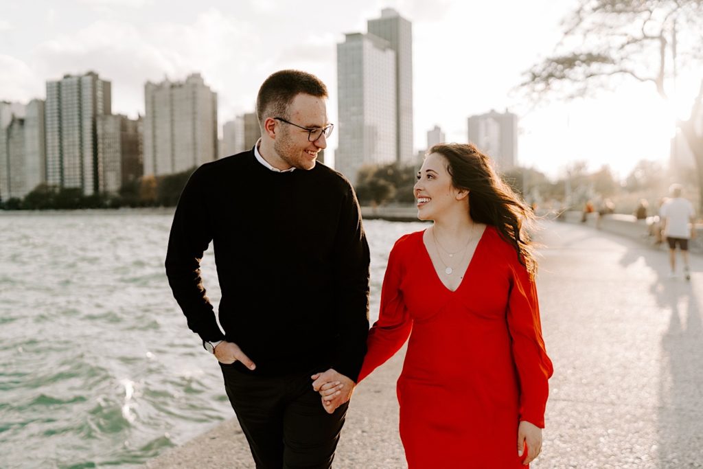 Couple walking together holding hands and smiling at one another on North Avenue Beach in front of the Chicago skyline and Lake Michigan during their engagement session