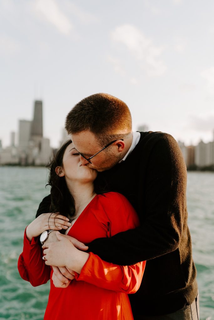 Man hugs fiancée from behind and kisses her on the lips at North Avenue Beach in front of Lake Michigan and the Chicago skyline