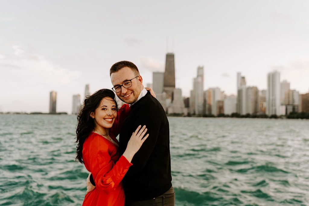 Couple embrace and smile at the camera together at North Avenue Beach in front of Lake Michigan and the Chicago skyline during their engagement session