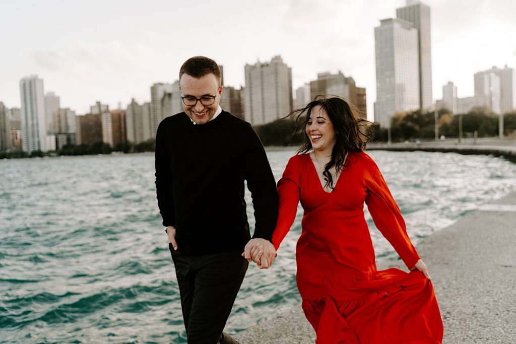 Couple walking together holding hands and smiling at North Avenue Beach in front of Lake Michigan and the Chicago skyline during their engagement session