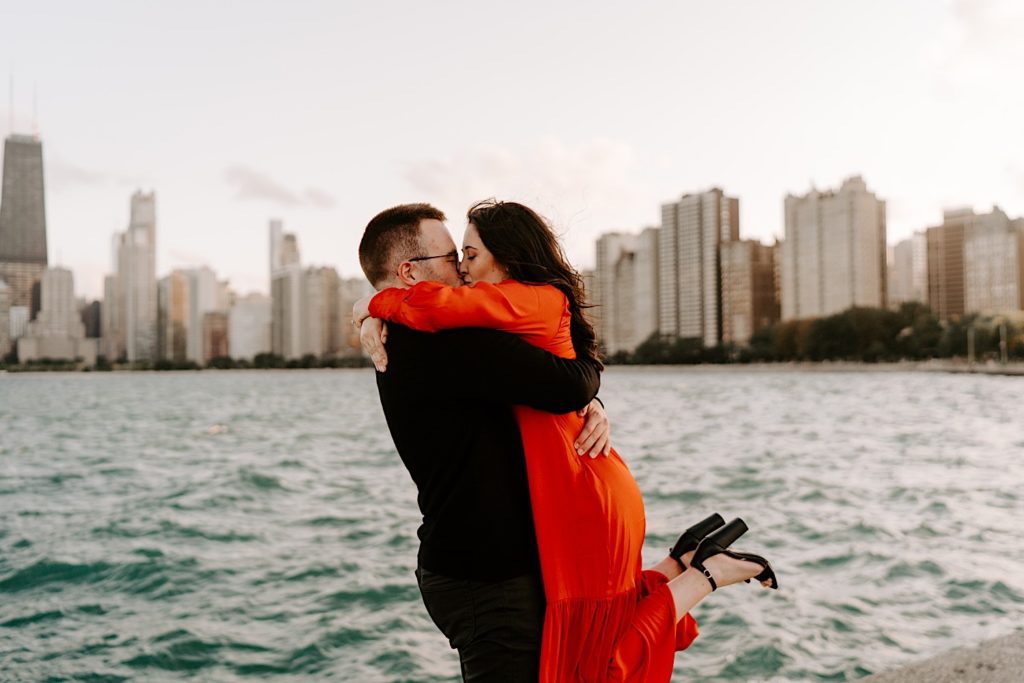Man hugs and lifts his fiancée while they kiss at North Avenue Beach with the Chicago skyline and Lake Michigan behind them during their engagement session