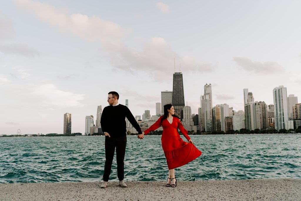Couple standing side by side holding hands and looking opposite directions at North Avenue Beach in front of the Chicago skyline and Lake Michigan during their engagement session