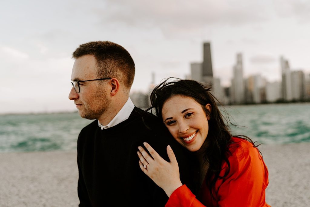 Woman holds her fiance from behind and smiles at the camera at North Avenue Beach with the Chicago skyline and Lake Michigan behind them during their engagement session