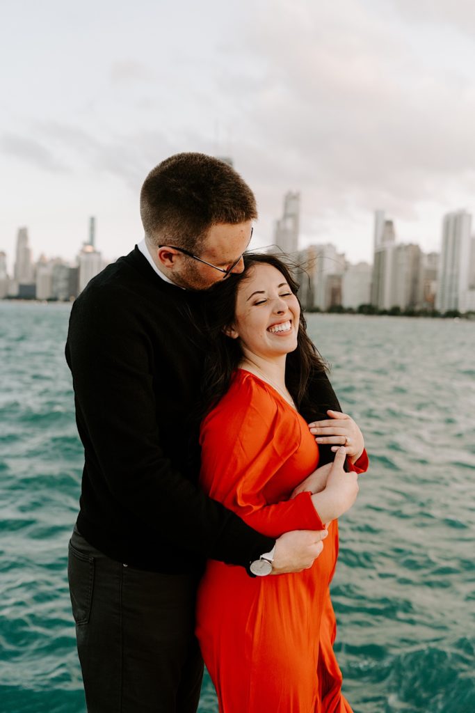 Man kisses his fiancée's head while holding her from behind as she smiles at North Avenue Beach with Lake Michigan and the Chicago skyline behind them