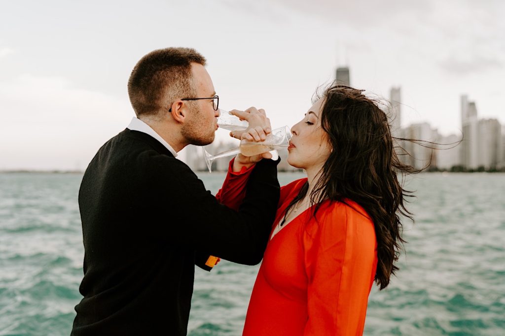 Couple lock arms and drink champagne together to celebrate their engagement at North Avenue Beach with Lake Michigan and the Chicago skyline in the background