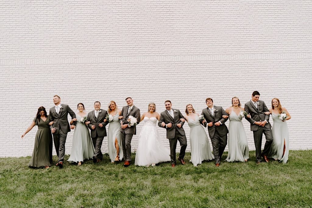 Wedding party linking arms together in front of white brick wall