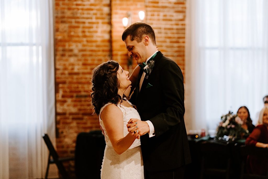 Newly weds smile at one another during their first dance at their receptions space while guests watch