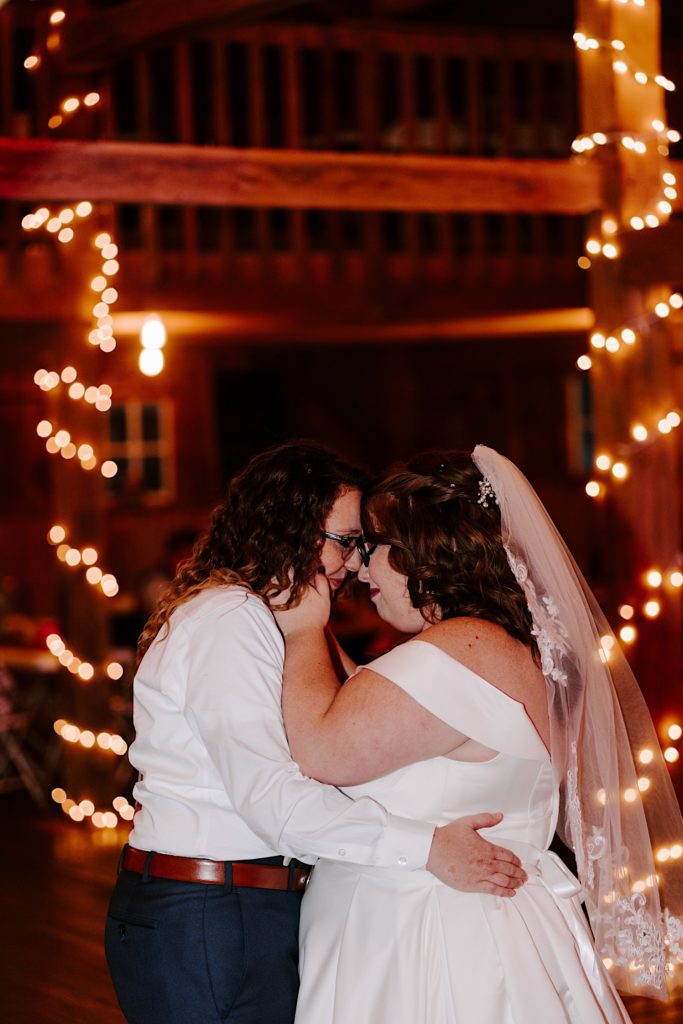 Brides share first dance together and touch foreheads together with string lights in the background of their reception space