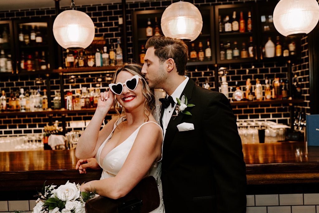 Bride wearing heart shaped sunglasses in front of bar being kissed on side of the head by groom