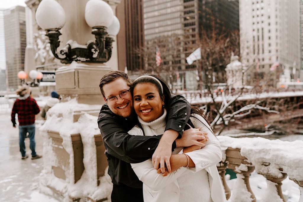 Fiance holding bride to be from behind smiling into camera during riverwalk engagement session