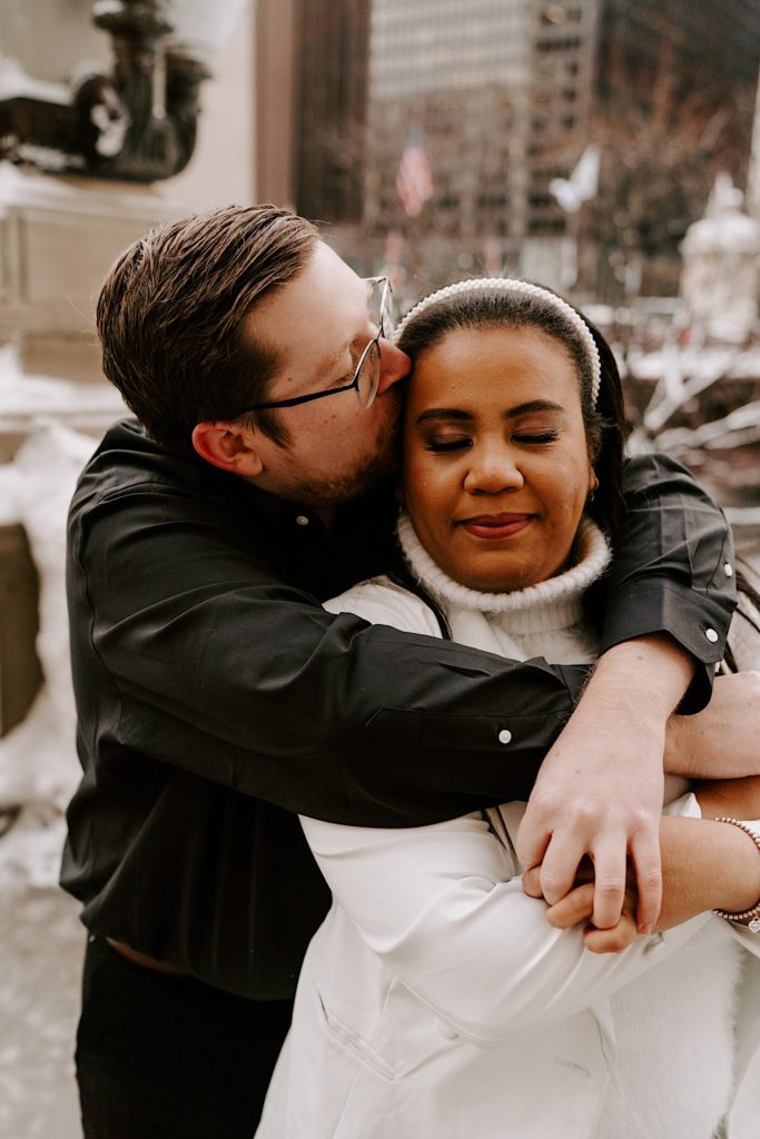 Fiance holding bride to be from behind and kissing her head in winter riverwalk engagement session