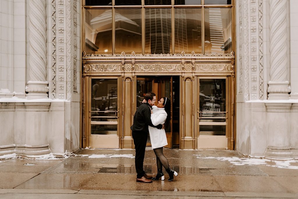 Engaged couple standing in front of golden bronze doors embracing one another