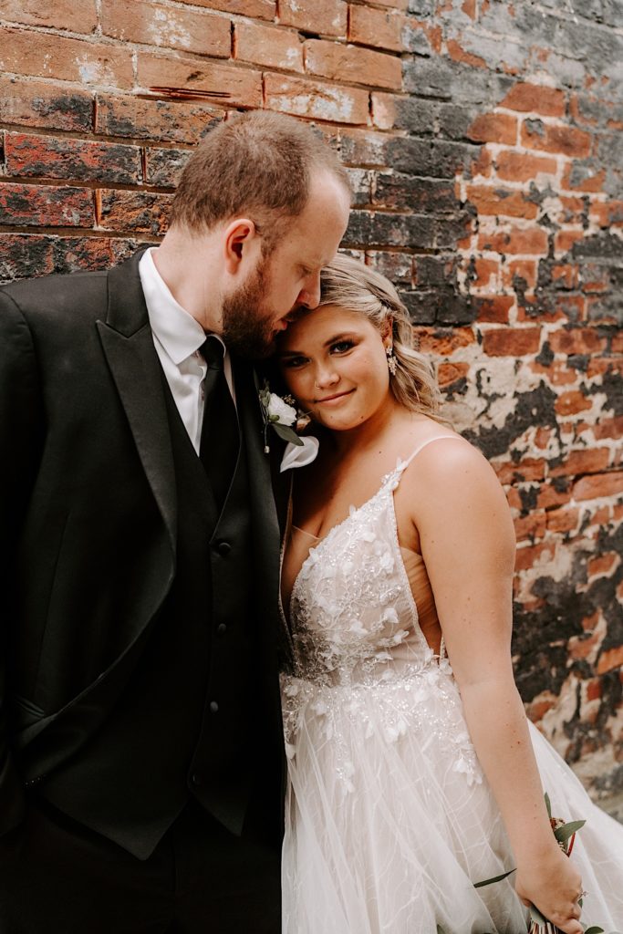Groom kisses brides forehead while leaning against brick wall