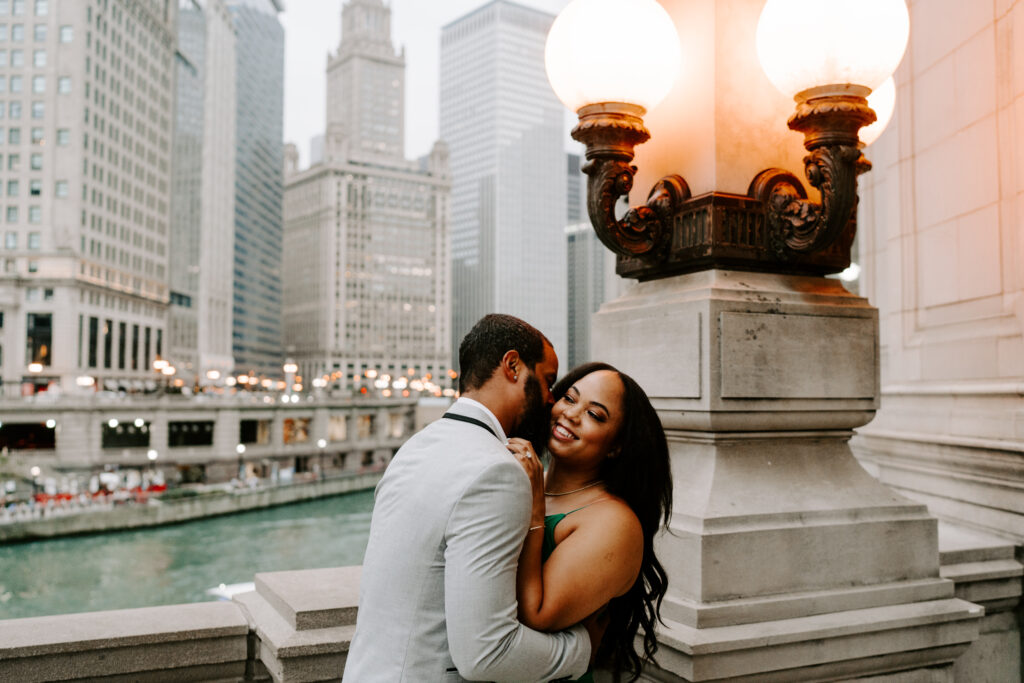 A couple kisses overlooking the Chicago river while taking their engagement photos.  She is wearing a green dress and has long black hair while he is wearing a grey suit.