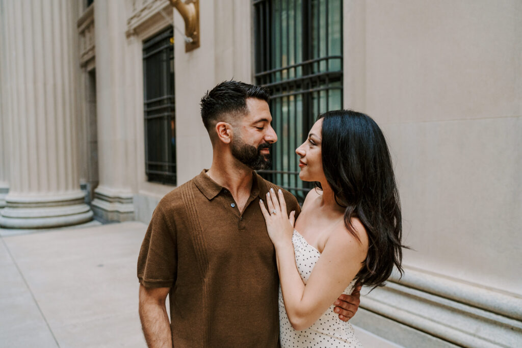 An engaged couple looks at one another during their engagement session at the Art Institute in Chicago.  The woman is wearing a polk-a-dot dress and the man is wearing a knit brown polo.