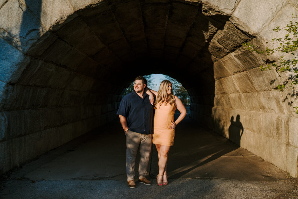 An engaged couple stands under an arch at the Lincoln Park Boardwalks, the woman is in an orange summer dress and the man is wearing a blue short sleeved button up