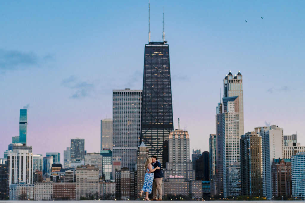 A couple kisses during sunset at North Avenue beach in Chicago while the sun sets, the sky is shades of pink and blue and you can see the chicago skyline