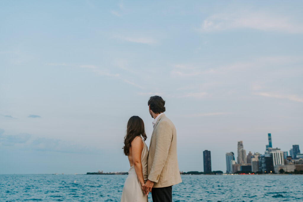 A couple stands holding hands while facing each other looking out over Lake Michigan at North Avenue Beach during their engagement session.