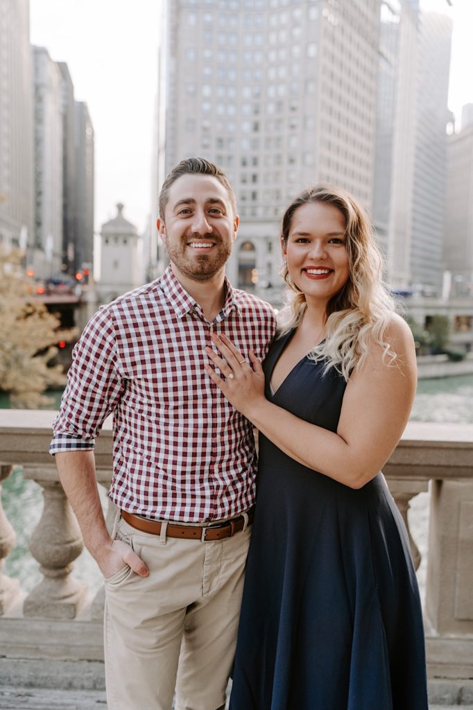 Couple smiles at the camera during their engagement session wearing a red plaid button up and a navy blue dress.