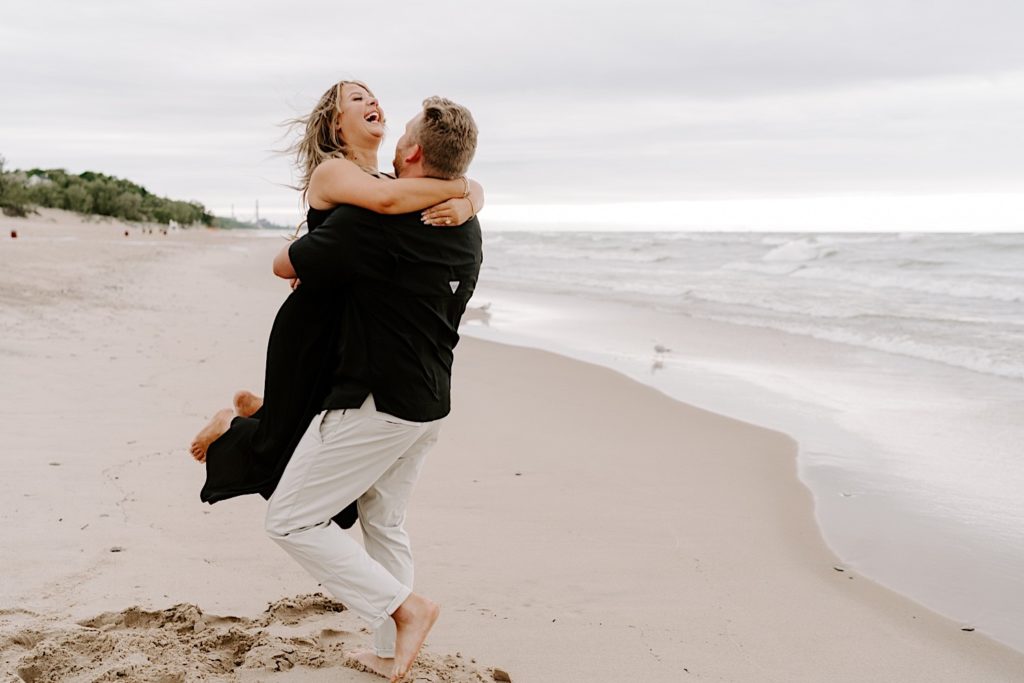 Groom picks up bride during their engagement session on the beach on Lake Michigan.