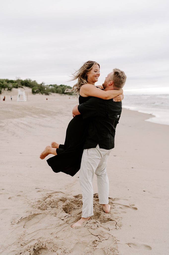 Couple holds each other on the beach.  The bride is wearing a black dress while the groom wears khakis and a black shirt