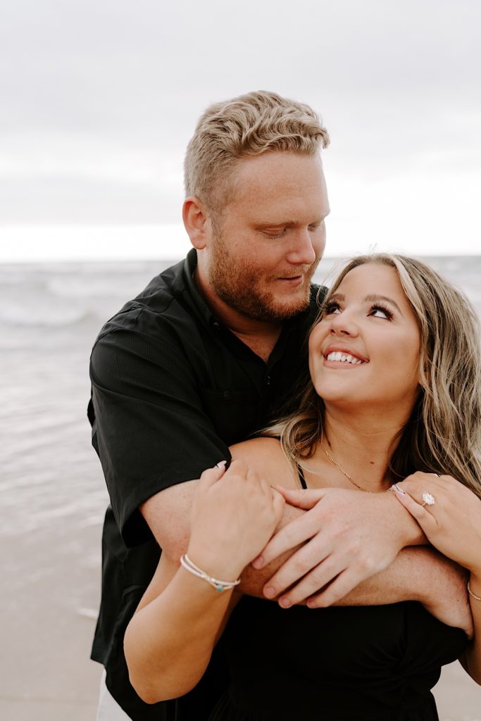 Bride looks up at groom while hugging during their beach engagement session
