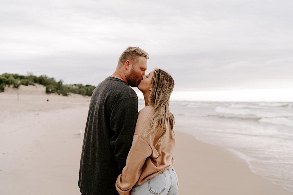 Couple kisses in beige and black sweatshirts during their cozy beach engagement session.