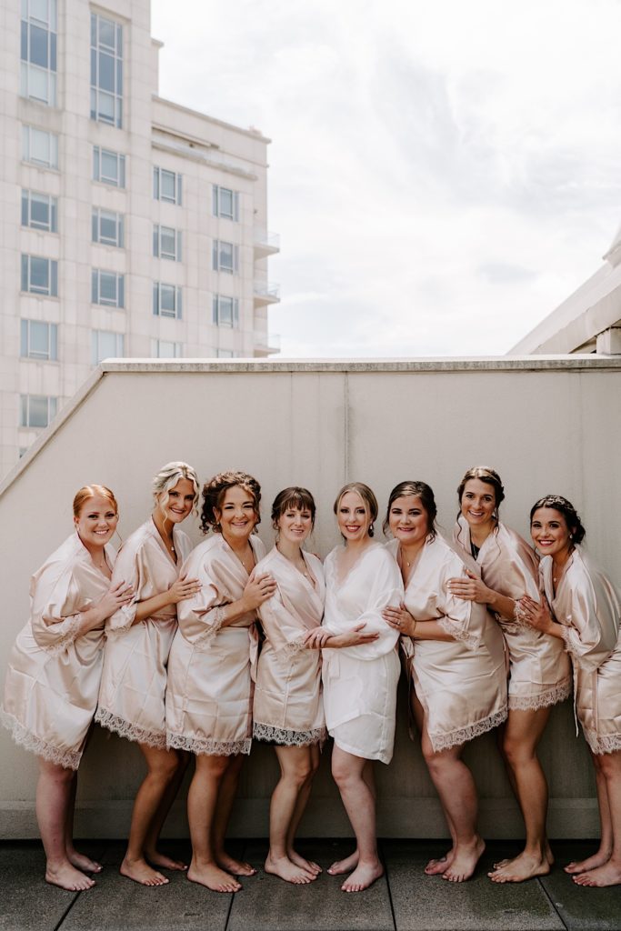 Bride stands with her wedding party in matching blush robes with a lace trim at the Embassy suites in Downtown Indy.