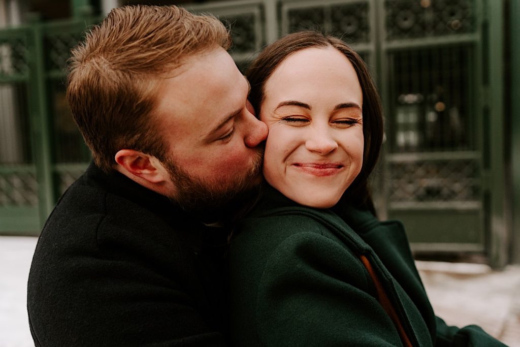 Future groom kisses future bride on the cheek during their winter engagement session.