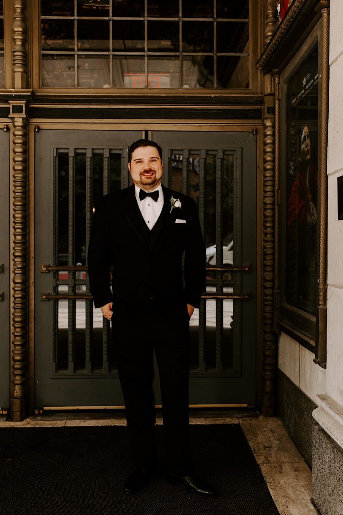 Groom stands outside of the the Indianapolis theater smiling at the camera with a white rose boutonnière