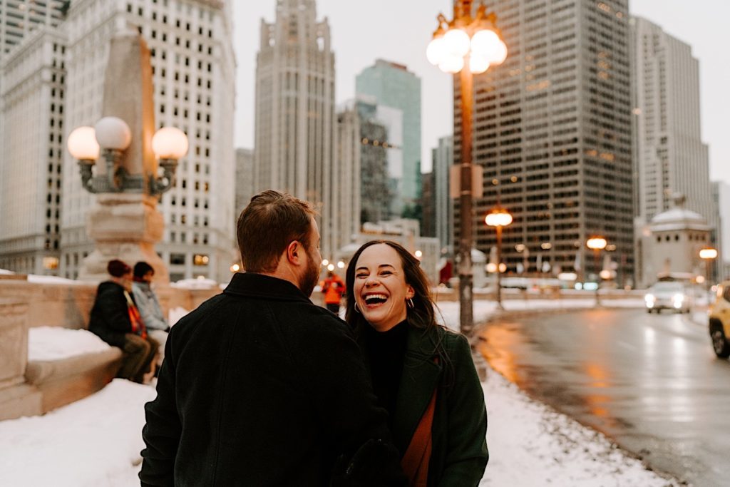 engaged couple laughs while walking along the Chicago River walk on a winter night.