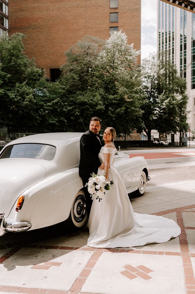 Bride and groom leans up against a white classic car for wedding portraits outside the Indiana Repertory Theatre