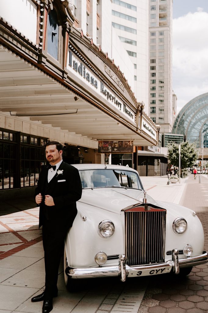 Groom leans up against a white 56 Rolls Royce outside the Indiana Repertory Theatre