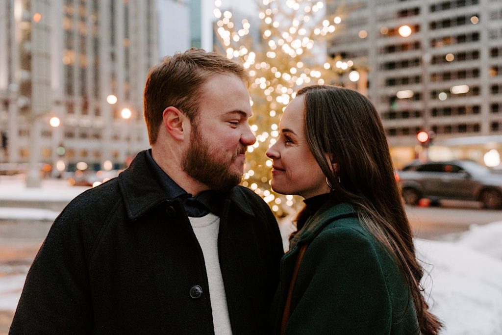 Couple brings their faces close together while standing in front of a Christmas tree for their winter Chicago engagement session.