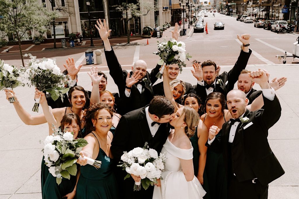 Bride and groom stand with their wedding party in Monument circle Indianapolis with the wedding party cheering and the couple kissing.