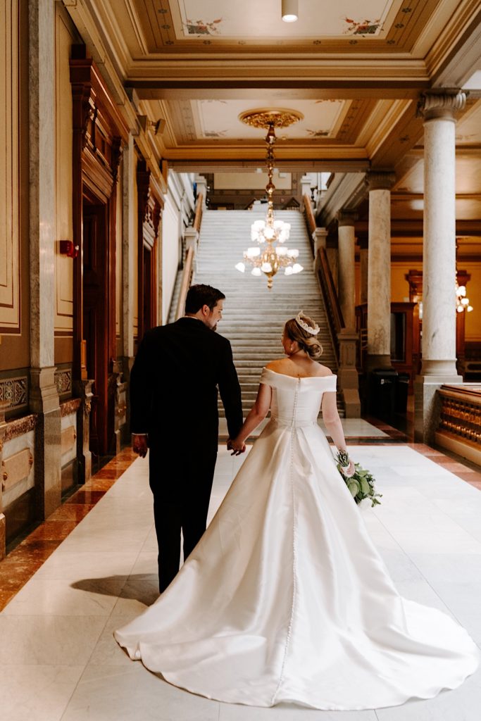 Bride wearing a pearl tiara walks with her groom towards a staircase at the Indiana State House before their wedding ceremony.
