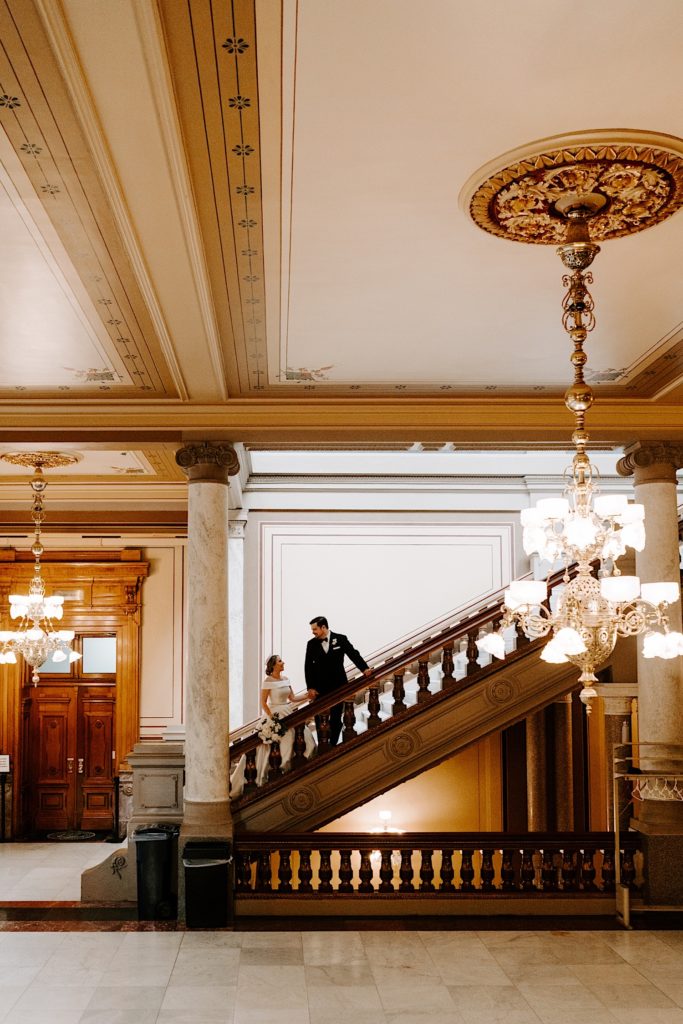 Bride and groom look at one another while standing on an ornate staircase at the Indianapolis State House prior to their wedding ceremony.