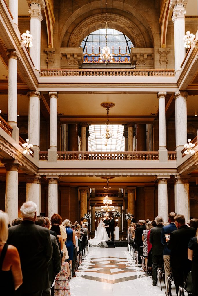 Bride and groom exchange vows standing in front of a crowd of 100 wedding guests at the Indiana State House for their wedding ceremony.