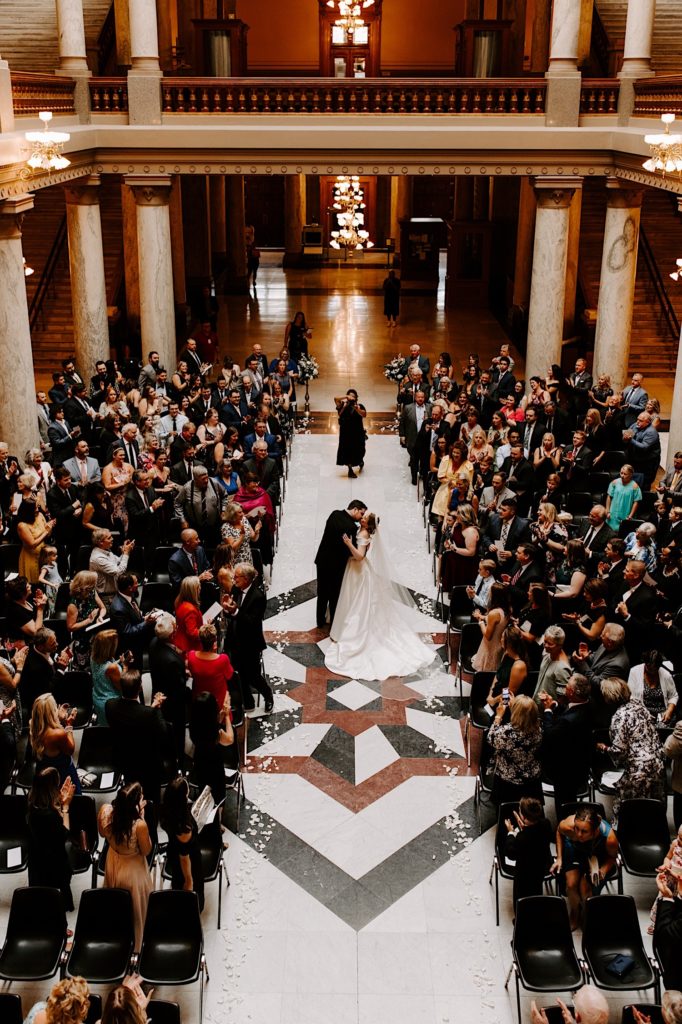 A top view photo looking over a balcony of a bride and groom kissing halfway down the aisle surrounded by their 100+ wedding guests after their wedding ceremony at the Indiana State house