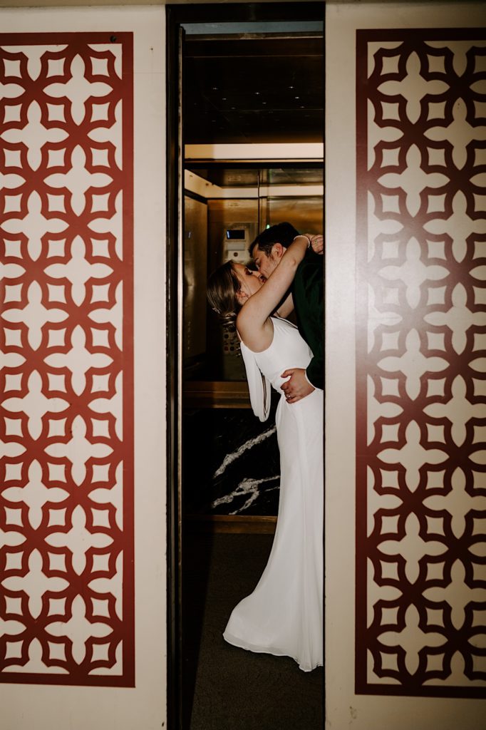 A black and white image of a bride and groom kissing in an elevator on their wedding day with the elevator door closing.