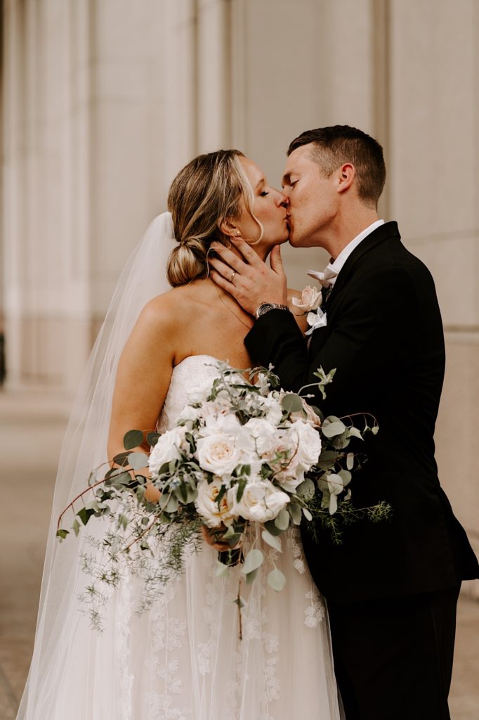 Bride holding an ivory rose bouquet while kissing her groom underneath the overhang at Union Station in Chicago.