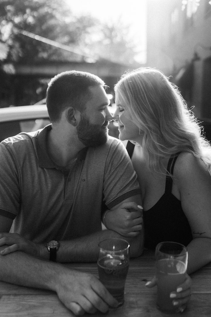 Couple sits outside at a brewery during their engagement session with their noses pressed together.