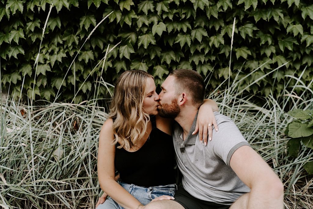 A couple kisses during their engagement session in front of a wall covered in ivy in Ravenswood Chicago